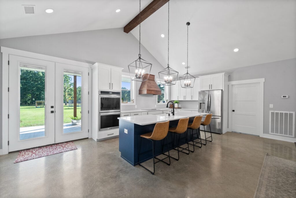 Bright, spacious kitchen with white cabinets, stainless steel appliances, and a large island with brown leather chairs. Open-concept design showcases high ceilings and views of the outdoors.